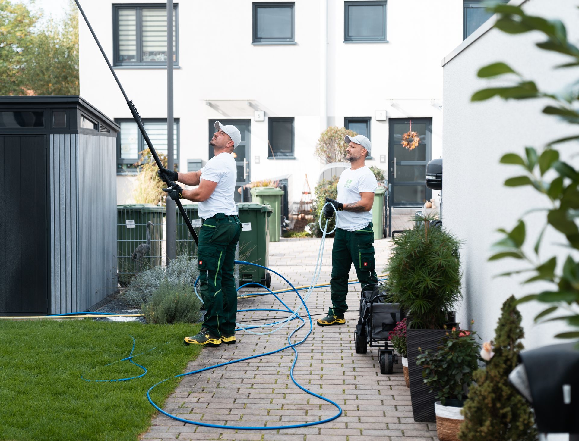 Zwei Mitarbeiter reinigen mit Teleskoplanze und Schlauch die Fassade eines Reihenhauses im Innenhof.