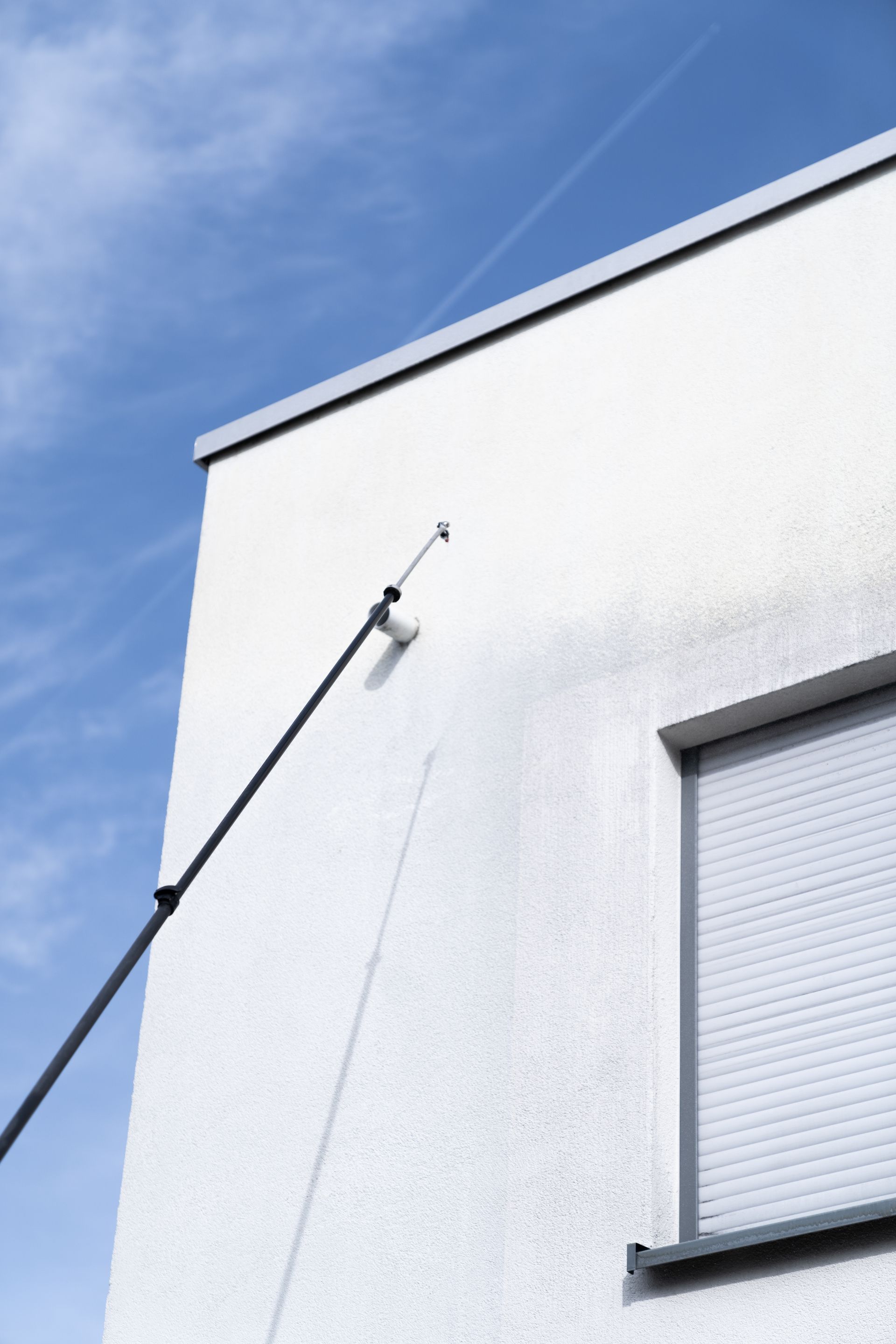 Weiße Gebäudekante vor blauem Himmel, im Anschnitt die Teleskoplanze beim Reinigen.